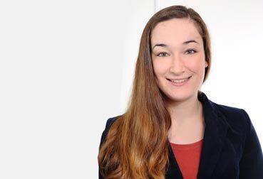 Lächelnde Frau mit langen Haaren im marineblauen Blazer und rotem Oberteil vor hellem Hintergrund.