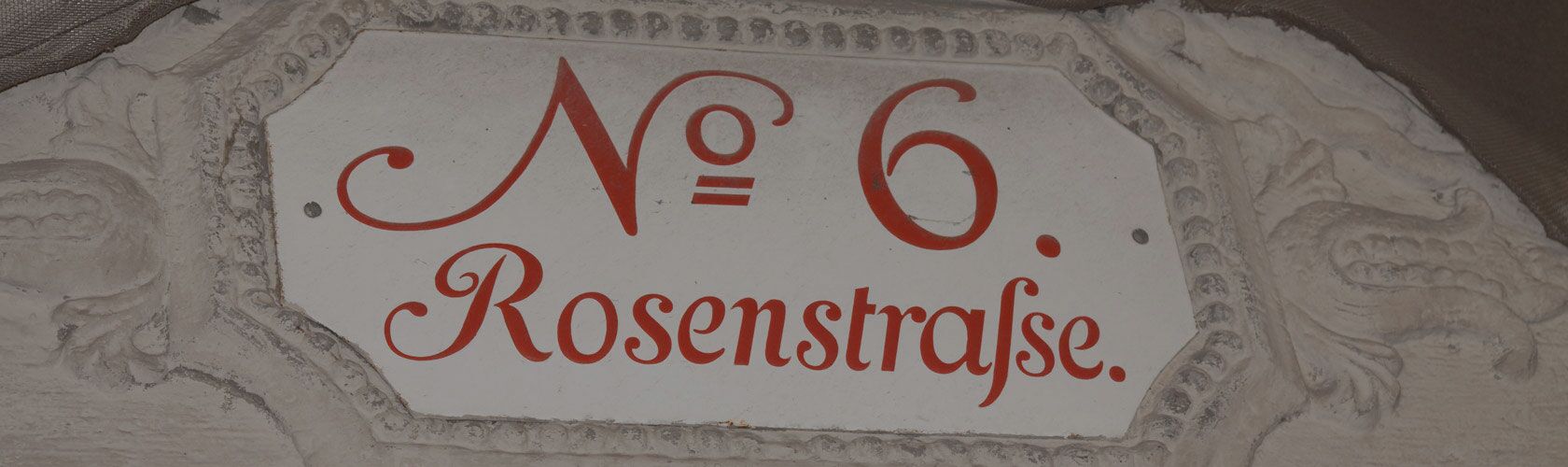 Hausnummer 6 in der Rosenstraße, kunstvoll auf einem weißen Schild mit roten Buchstaben und dekorativem Rahmen gestaltet.