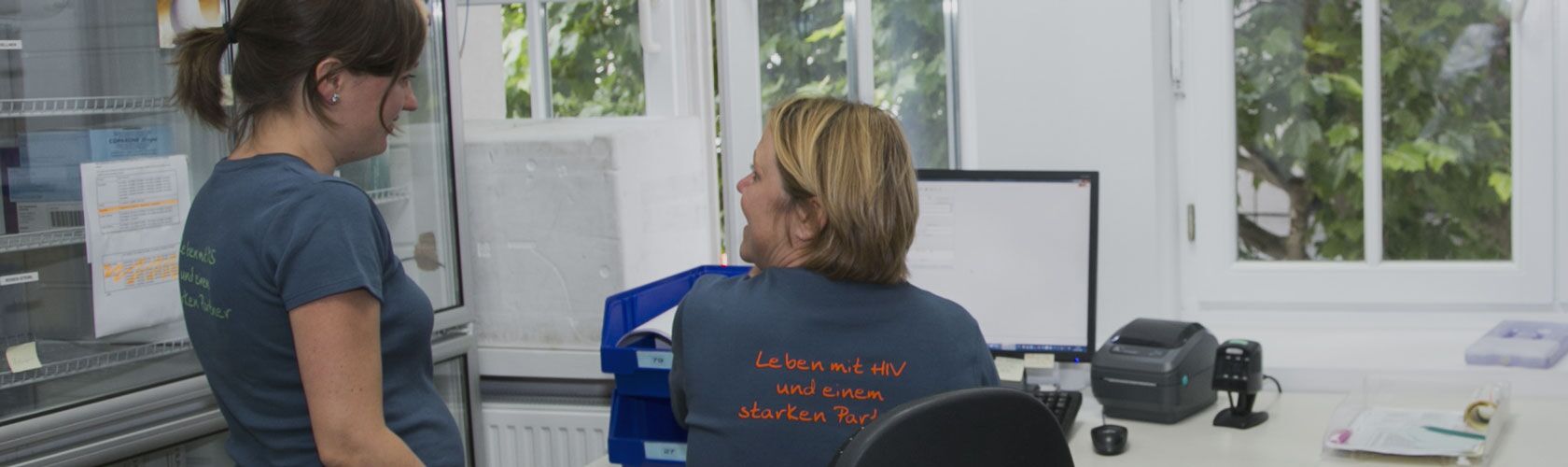 Zwei Frauen in einer Apotheke diskutieren neben einem Computer. Eine trägt ein T-Shirt mit einem motivierenden Spruch.