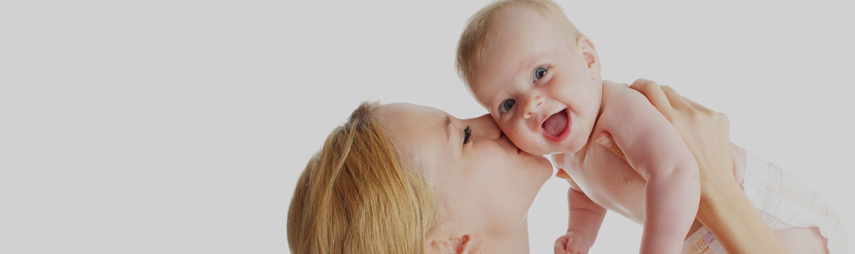 Une mère blonde embrasse tendrement son bébé souriant, en le tenant dans ses bras sur fond clair.