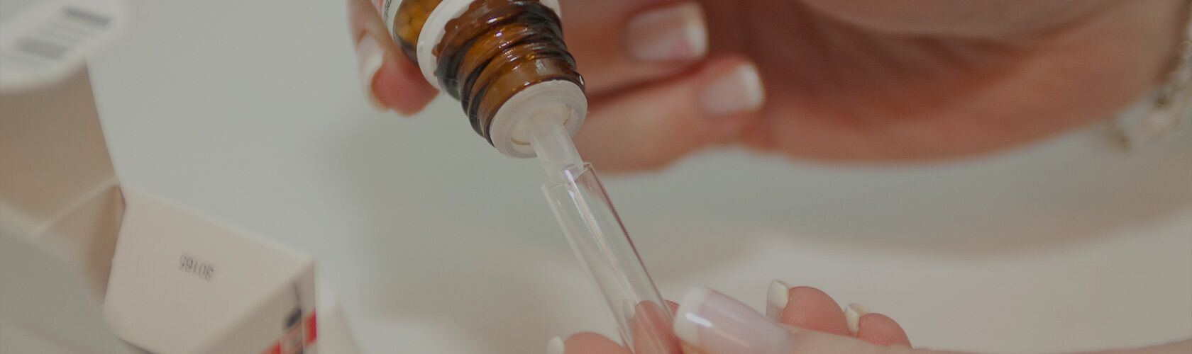 Main en train de préparer un remède homéopathique avec une pipette et un flacon brun sur une table blanche.