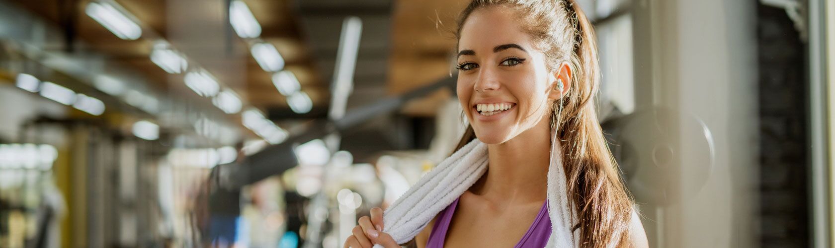 Lächelnde Frau in Sportkleidung mit Handtuch über den Schultern im hellen Fitnessstudio, verkörpert Gesundheitsvorteile.