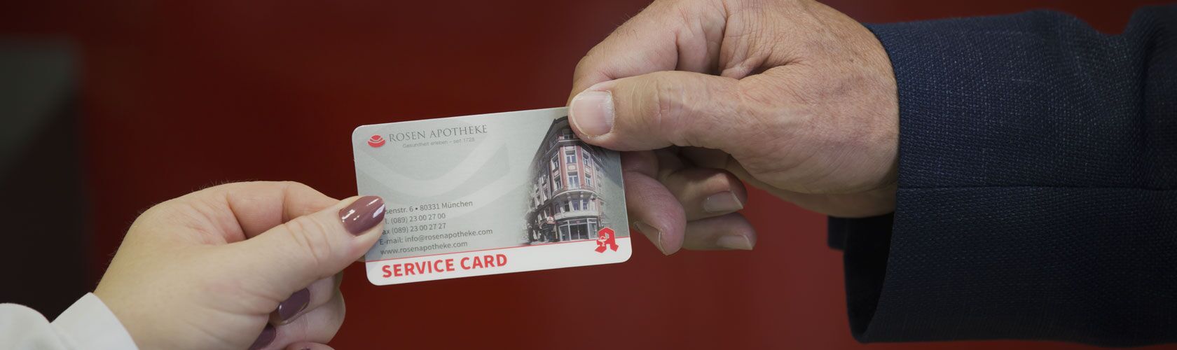 Eine Hand überreicht eine Servicekarte der Rosen Apotheke an eine andere Hand vor einem roten Hintergrund.